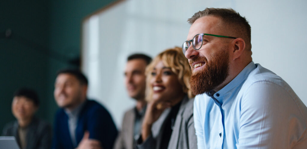 Smiling businessman and unrecognizable multi-ethnic employees sitting in a row at their company desk and listening to their boss.